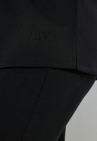 Vêtements de sport noirs avec une texture lisse, arborant un logo "MV" en accents étincelants près de l'ourlet. Coupe ajustée.