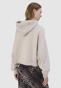Vrouw draagt een beige cropped hoodie met "California" geborduurd op de capuchon en "Los Angeles" op de zoom aan de achterkant, gecombineerd met een leopardprint rok.