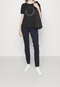 Camiseta de algodón negra con gráfico plateado circular, combinada con pantalones ajustados azul marino y zapatillas blancas. El bolso es negro y acolchado.