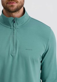 Lichtgroene quarter-zip pullover van zacht materiaal. Heeft een gestikte kraag en een subtiele logo op de borst. Glad textuur.