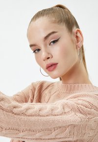 Różowy sweter z dzianiny o teksturowanym wzorze w warkocze. Modelka ma srebrne kolczyki w kształcie kół oraz koński ogon z boku. Gładka skóra i makijaż podkreślają rysy.