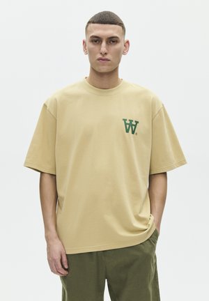 Ung mand med kort hår iført en beige oversized t-shirt med grønt "W" logo og olivengrønne bukser, stående med den ene hånd i lommen.