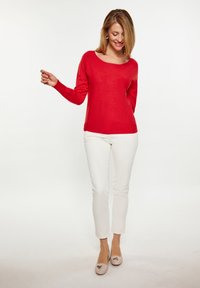 Roter langärmeliger Pullover mit rundem Ausschnitt, kombiniert mit weißen Slim-Fit-Hosen. Die Schuhe sind hellbeige mit einem dekorativen Akzent. Einfacher Hintergrund.
