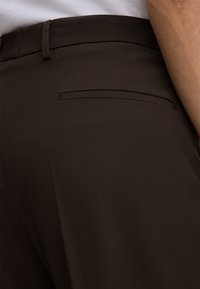 Dunkelbraune Hose mit glatter Textur, schlanker Passform und einer einzelnen Leistentasche hinten. Minimalistisches Design ohne sichtbare Beschläge.