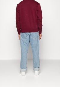 Felpa color marrone con polsini a costine, abbinata a jeans in denim azzurro chiaro. I jeans hanno una vestibilità rilassata e orli risvoltati, indossati con sneakers bianche.