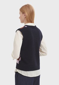 Marinblå ärmlös stickad väst med flerfärgade geometriska mönster, texturerad ullblandning, lager över en ljusfärgad skjorta med knappar.