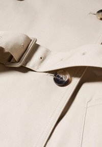 Gros plan sur un tissu beige avec une ceinture assortie comportant des boutonnières cousues et un bouton translucide à quatre trous cousu sur le matériau.