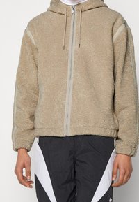 Beige fleecjacka med dragkedja fram, justerbar huva och textilstruktur. Har beige sidosträck och elastiska ärmslut.