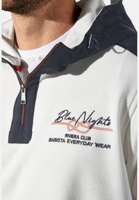 Weißer Hoodie mit einem marineblauen Kragen, ausgestattet mit einem halben Reißverschluss, besticktem "Blue Nights"-Logo und roten Akzenten bei der Naht.