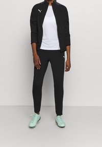 Zwarte lichte Puma-jas met opstaande kraag en wit logo; wit aansluitend shirt; zwarte sportbroek; mintgroene schoenen met veters.