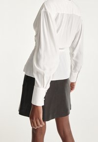 Vit blus med voluminösa ärmar, samlad vid midjan och med knapppynt vid ärmsluten. Bärs med en mörkgrå kjol.