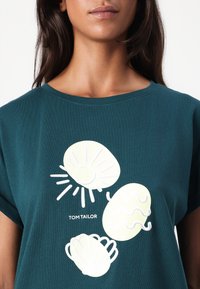 Grønn bomull T-skjorte med hvit grafikk av en sol, skjell og bølgete eggformer. Merket leser "TOM TAILOR." Korte ermer, avslappet passform.