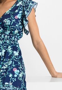 Vestido floral azul marino con cintura fruncida, mangas volantes y patrones botánicos en azul claro y rosa sobre un fondo oscuro.