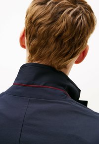 Giacca blu navy con superficie testurizzata, caratterizzata da un colletto alla coreana e un accento color vinaccia lungo il bordo del colletto. Vista posteriore del capo.