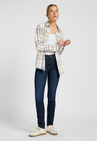 Rutig flanellskjorta i krämfärg med rosa och blå accenter, tillsammans med mörkblå smala jeans och vita sneakers med beige accenter.