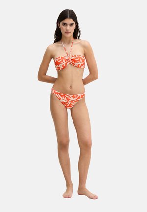 Young woman standing barefoot, wearing orange and white leaf-patterned bandeau bikini with halter strap, against plain light background.