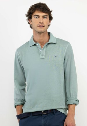 Hombre con cabello corto y rizado, vestido con una camisa polo de manga larga azul claro y pantalones azul marino, de pie sobre un fondo blanco liso.