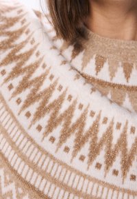Nahaufnahme eines beigen und weißen gemusterten Wollpullovers mit geometrischen Designs und subtilen schimmernden Fäden, getragen von einer Person mit braunen Haaren.
