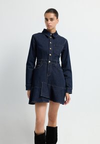 Robe chemise en denim foncé avec un col, une fermeture à boutons sur le devant et une poche patch. Taille cintrée et ourlet évasé. Présente des coutures contrastantes et des poches.