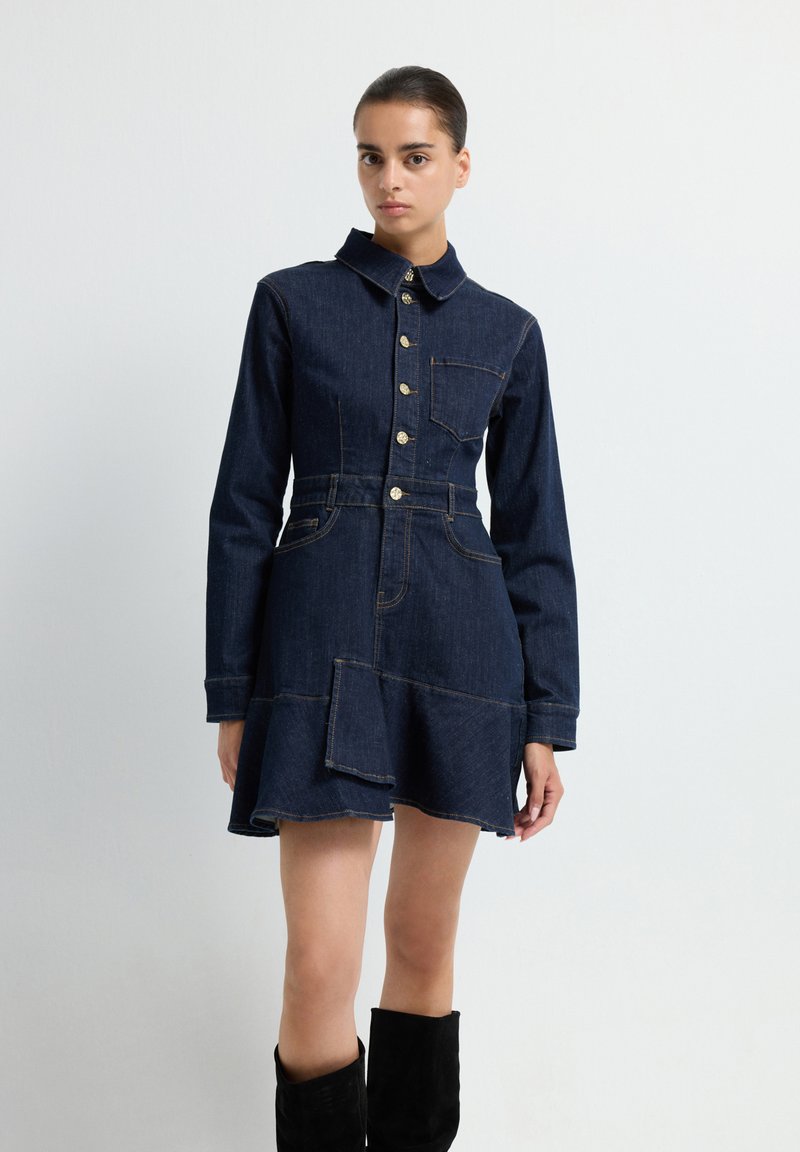 Robe chemise en denim foncé avec un col, une fermeture à boutons sur le devant et une poche patch. Taille cintrée et ourlet évasé. Présente des coutures contrastantes et des poches.