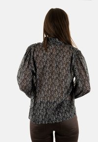 Blusa negra transparente con mangas largas abullonadas, intrincado patrón de paisley en azul y blanco, cuello alto y detalle de fruncido en la parte trasera.