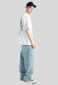 Biała, oversize'owa koszulka z graficznym wzorem, jasnoniebieskie luźne dżinsy, czapka w kolorze czarnym oraz białe trampki z niebieskimi akcentami, zaprezentowane na szarym tle.