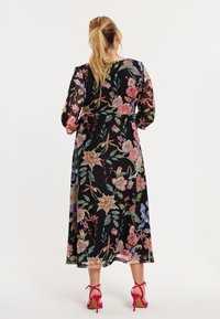 Zwarte maxi-jurk met bloemenprint, kleurrijke patronen en lange mouwen. Voorzien van een taillestrik en een vloeiende silhouet. Gecombineerd met roze sandalen met hoge hakken.