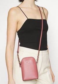 Pink pebbled leather crossbody bag with a rounded shape, chain strap, and silver logo accent. Worn over a black top and cream pants.