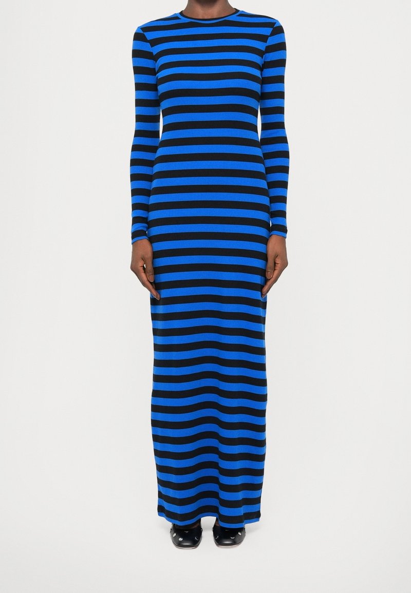 Femme portant une longue robe moulante à rayures horizontales noires et bleues, debout avec les mains détendues le long du corps devant un fond uni.
