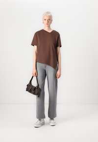 Brun, asymmetrisk t-skjorte i bomull, kombinert med lysegrå rette jeans. Modellen holder en liten, mørkebrun håndveske og har på seg hvite sneakers.