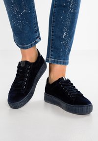 Námořnické sametové tenisky s plochou gumovou podrážkou, šněrovacím designem a texturovanými detaily. Nošeny s výrazně opotřebovanými modrými džíny.