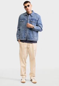Giacca di jeans azzurro chiaro con vestibilità rilassata, chiusura a bottoni frontale, tasche doppie sul petto, abbinata a pantaloni beige e sneakers casual.
