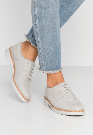 Lys grå ruskindssko med snørebåndsdesign, hvide gummisåler og kontrasterende korkdetaljer. Båret med slidte denimjeans.