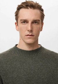 Pull tricoté vert foncé avec un col rond côtelé et un tissu texturé, porté par une personne aux cheveux courts et châtain clair, au regard neutre.