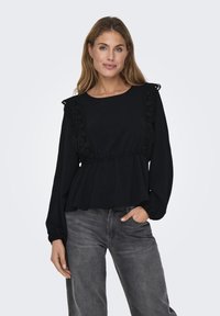 Svart langermet topp med rynket midje, prydet med blonder langs skuldrene, laget av et mykt, teksturert stoff. Kombinert med grå jeans.
