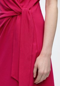 Gros plan sur le bras d’une personne à côté d’une robe rose vif avec un grand détail en tissu noué à la taille.