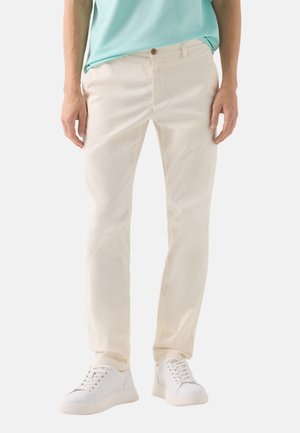 Mand iført lysebeige slim-fit bukser, hvide sneakers og en lyseblå skjorte, stående mod en hvid baggrund.