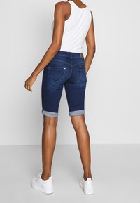 Person som bär mörkblå uppvikta denimshorts, vit linne och vita sneakers, står mot en ljus, enfärgad bakgrund vänd bort.