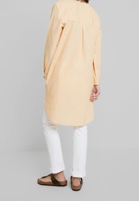 Langärmelig shirt in lichtoranje, met verticale strepen, een plooi op de rug, een gebogen zoom, gecombineerd met witte broek en bruine sandalen.