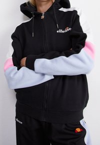 Personne portant un sweat à capuche noir zippé et un pantalon assorti avec des blocs de couleur blanc, rose et bleu clair, arborant le logo "ellesse" sur la poitrine et la jambe.