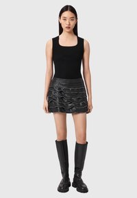 Γυναίκα που φοράει μαύρο αμάνικο ριμπ τοπ, μίνι φούστα από μαύρο δέρμα με οριζόντιες βολάν και μαύρες μπότες μέχρι το γόνατο, στέκεται μπροστά σε γκρίζο φόντο.