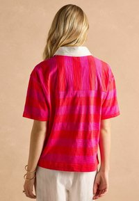 Krátky rukáv pruhovaný top v jasne ružových a červených odtieňoch s bielym golierom a textúrovanou tkaninou. Má voľný strih a bočné rázpory.
