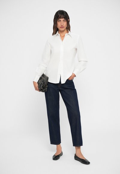 Camicia bianca a maniche lunghe con colletto classico, abbinata a jeans dritti blu scuro e scarpe nere basse, mentre tiene una borsa strutturata.