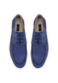 Des chaussures en daim bleu avec une texture lisse, présentant un design basse, un laçage à l'avant et des semelles en caoutchouc blanches. Étiquette de marque visible à l'intérieur.