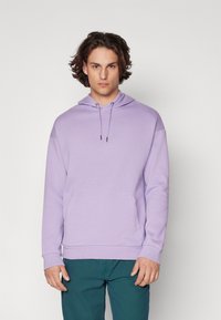 Felpa viola chiaro con tasca a marsupio e cappuccio con cordino. Presenta polsini e orlo a costine, realizzata in tessuto morbido. Il modello indossa pantaloni turchesi.