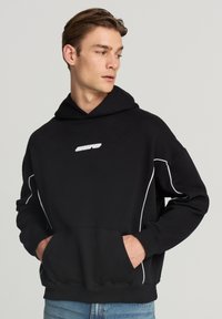 Uomo che indossa un hoodie nero con piping bianco e logo, con le mani nella tasca anteriore, che guarda di lato su uno sfondo neutro.