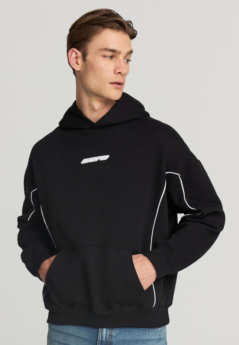 Uomo che indossa un hoodie nero con piping bianco e logo, con le mani nella tasca anteriore, che guarda di lato su uno sfondo neutro.