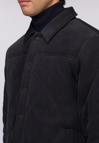 Kvadret mörkgrå kordjacka med krage, tryckknappar och ett diagonalt sydd mönster på framsidan.