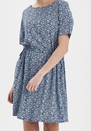 Femme portant une robe bleu marine à manches courtes, longueur genou, avec un petit motif floral blanc et une ceinture nouée à la taille.