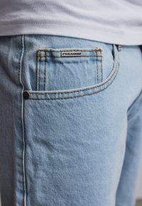 Person trägt hellblaue Jeans mit sichtbarer Fronttasche und weißem Bund, Markenlabel "PEGADOR" über der Tasche.